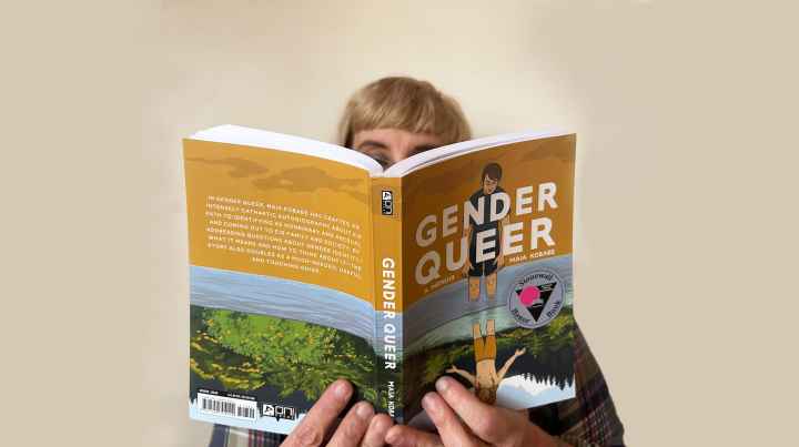 A white person with blond hair and glasses reads a copy of "Gender Queer" against a solid white backdrop