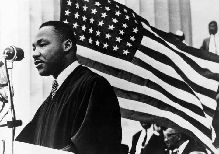 Black and White Photo shows Dr. Martin Luther King Jr. in front of an American flag