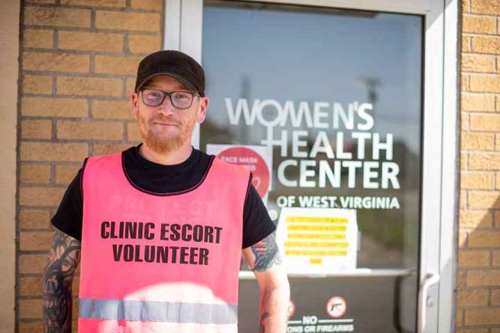 Rusty Williams wears a pink vest reading Clinic Escort Volunteer in front of the entrance to the Women's Health Center of West Virginia