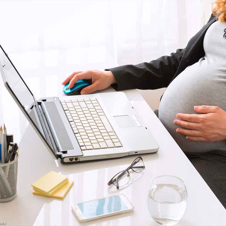 A pregnant worker touches their belly while performing work duties on a laptop
