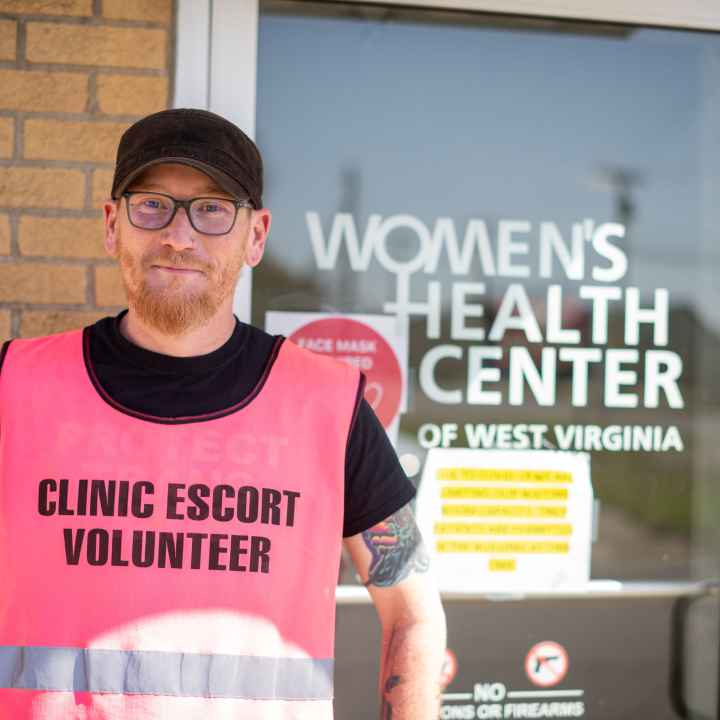Rusty Williams wears a pink vest reading Clinic Escort Volunteer in front of the entrance to the Women's Health Center of West Virginia