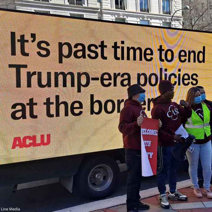 Protestors stand in front of a digital sign that says "It's past time to end Trump-era policies at the border"